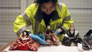 Someone wearing a hi-vis vest and blue latex gloves is carefully arranging the fringe on a Japanese doll placed on a white table. There are four dolls visible. They are seated and about 6 inches high. They are all dressed in sumptuous Japanese clothing, with embroidery and embellishments.